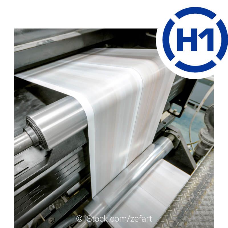 Blick in eine laufende Rollendruckmaschine. Das frisch bedruckte Papier wird über mehrere Rollen transportiert. Rechts oben ist das H1-Logo abgebildet.